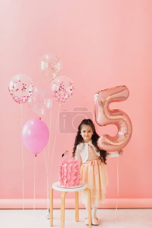 Photo for Adorable little girl in a pink dress posing next to a flamingo birthday cake and celebrating her fifth birthday on a pink background - Royalty Free Image