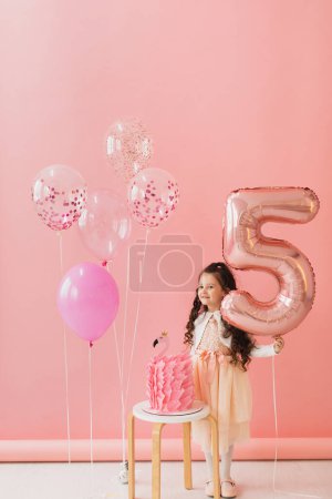 Foto de Adorable niña en un vestido rosa posando junto a un pastel de cumpleaños flamenco y celebrando su quinto cumpleaños sobre un fondo rosa - Imagen libre de derechos