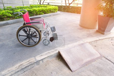 Silla de ruedas roja vacía estacionada en el hospital cerca de la placa de diamante rampa de apoyo para personas con discapacidad.