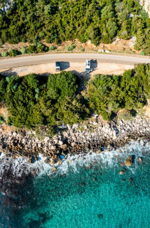 Photo for A top down view of two camper vans parked on a narrow coastal road in the forest with turquoise water and rocky shore - Royalty Free Image