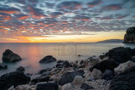 Téléchargez les photos : Un lever de soleil coloré à Cala Gonone plage avec des roches noires et blanches et des rochers au premier plan - en image libre de droit