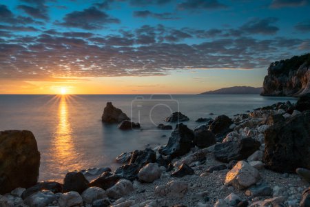 Téléchargez les photos : Un lever de soleil coloré à Cala Gonone avec des roches et des rochers noirs et blancs et un coup de soleil - en image libre de droit