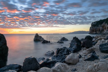 Téléchargez les photos : Un lever de soleil coloré à Cala Gonone plage avec des roches noires et blanches et des rochers au premier plan - en image libre de droit