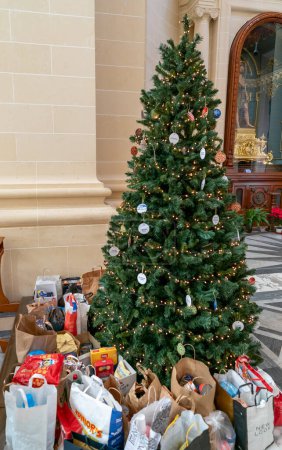 Téléchargez les photos : Xewkija, Malte - 20 décembre, 2023 : Arbre de Noël à l'intérieur d'une église avec des dons de sacs d'épicerie et de nourriture autour - en image libre de droit