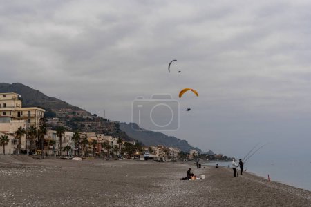 Foto de Letojanni, Italia - 29 de enero de 2023: pescadores en la playa de Letojanni con parapentes aterrizando detrás de ellos después de un vuelo desde las cimas de las montañas sicilianas - Imagen libre de derechos