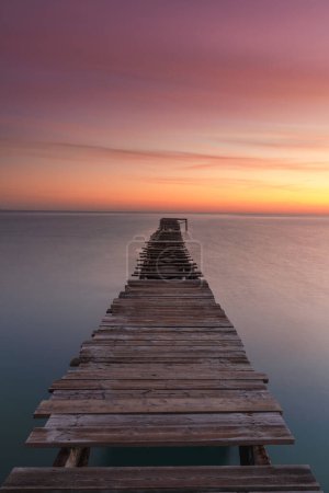 A vertical sunrise seascape with an old wooden dock leading out into the calm ocean waters