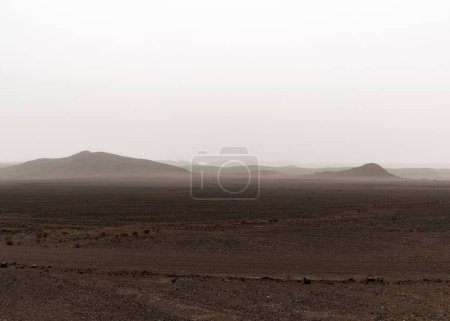 Eine Wüstenlandschaft mit trockenen Hügeln in der Ferne unter einem nebligen Sandsturmhimmel im Süden Marokkos