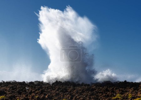 Huge wave crashing onto a rocky reef under a blue sky and creating an eruption of water