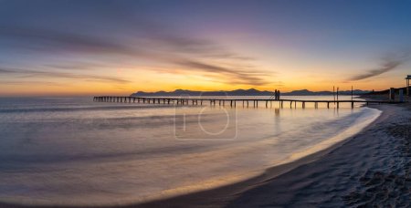 Peaceful sunrise seascape with an old wooden dock leading out into the calm ocean waters of Alcudia Bay