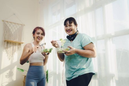 Foto de Concepto de fitness y comida saludable. Dos mujeres asiáticas tamaño del cuerpo diferente en ropa deportiva mirando cámara mientras sonríe disfrutar de la aptitud y feliz en comer alimentos saludables después de hacer ejercicio en casa juntos. - Imagen libre de derechos