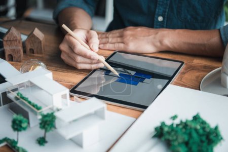 Photo for Architecture designer holds a pencil pointing to the 3D home office drawing on the tablet while thinking and considering detail in the building, Interior design and landscape around. - Royalty Free Image