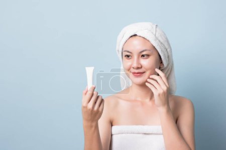 Téléchargez les photos : Belle jeune femme asiatique avec serviette de bain sur la tête souriante tenant de la crème blanche dans sa main, touchant son visage de peau éclatante avec l'espace de copie fond pastel bleu clair. - en image libre de droit