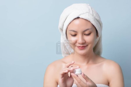 Téléchargez les photos : Portrait Asiatique jeune femme avec serviette de bain sur la tête en souriant tout en touchant le produit de crème de soins de la peau, lumineux visage de la peau avec espace de copie bleu clair fond pastel. - en image libre de droit