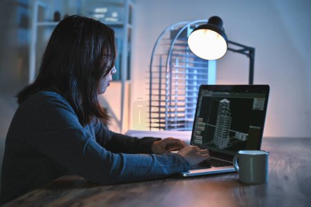 Asiatische Architektin oder Ingenieurin verwenden Laptop-Studie Detail und Konstruktion allein in Zeichnung in einem Büroraum in der Nacht mit Gebäudemodell auf Schreibtischen Büro.