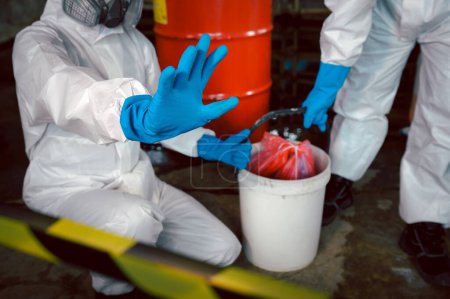 Oficiales especializados en seguridad química usan ropa protectora de riesgo químico levantados a mano diciendo precaución por derrames químicos mientras limpian y recuperan al llevar un cubo en parte de derrames químicos.
