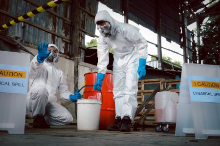 Oficiales especializados en seguridad química usan ropa protectora de riesgo químico levantados a mano diciendo precaución por derrames químicos mientras limpian y recuperan al llevar un cubo en parte de derrames químicos.