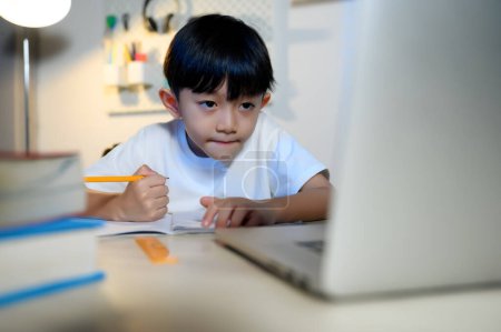 Téléchargez les photos : Asian Kid Boy se concentre sur les devoirs et l'apprentissage en ligne seul avec ordinateur portable dans la chambre à la maison, se concentrant sur l'apprentissage et faire des devoirs, étudier et réussir dans l'éducation des enfants asiatiques Concept. - en image libre de droit
