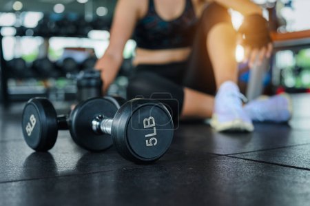 Photo for Dumbbell in the gym with woman blur while sit breaking relax after fitness exercise workout in background. Fitness exercise building muscle of women concept. - Royalty Free Image