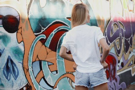 Foto de Chica rubia en camiseta blanca - camisa y pantalones cortos cerca de la pared de graffiti - Imagen libre de derechos