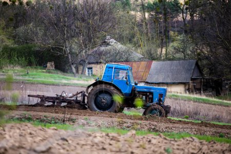 Photo for Old blue tractor with plow on field and cultivates soil. Preparing the soil for planting vegetables in spring. Agricultural machinery, field work. - Royalty Free Image