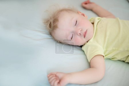 kleines Kind schläft mit ausgestreckten Armen. Mädchen mit blonden Haaren im gelben Body. Konzept gesunder Schlaf