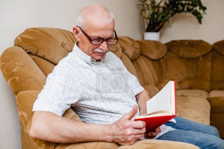 Téléchargez les photos : Homme âgé lisant un livre à la maison. Un homme d'affaires âgé fait un plan d'affaires assis à la maison dans une pièce sur le canapé. Organisation de l'entreprise par les gens depuis 65 ans - en image libre de droit