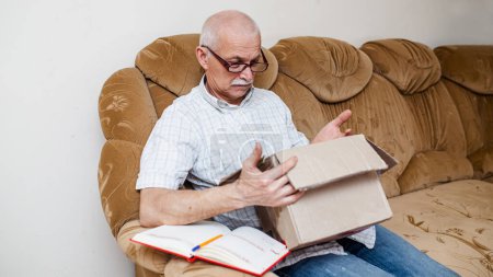 Téléchargez les photos : Un vieillard souriant a reçu un colis postal à la maison. Un grand-père surpris tient une boîte en carton ouverte. Commande du Service d'aide aux personnes âgées - en image libre de droit