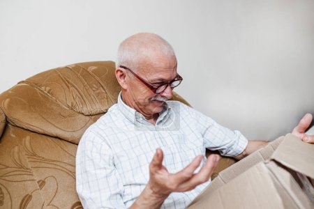 Téléchargez les photos : Un homme âgé est surpris par le contenu d'une boîte en carton faite d'un service de livraison rapide. Heureux propriétaire du colis envoyé par la société de service client en ligne. - en image libre de droit