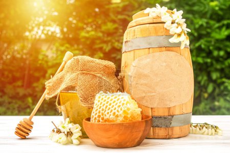 Photo for Text HONEY. Fresh liquid acacia honey harvest 2021 in a wooden barrel and a transparent jar with a canvas napkin on the lid. Branches of flowering white acacia - Royalty Free Image