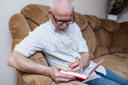 Téléchargez les photos : Le vieil homme prend des notes dans un planeur ou un carnet alors qu'il est assis à la maison sur le canapé. Pensionné travaille comme pigiste, fait des affaires après la fin de sa carrière - en image libre de droit