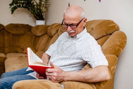 Téléchargez les photos : Homme âgé lisant un livre à la maison. Un homme d'affaires âgé fait un plan d'affaires assis à la maison dans une pièce sur le canapé. Organisation de l'entreprise par les gens depuis 65 ans - en image libre de droit