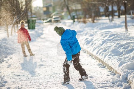 Kinder spielen auf der schneebedeckten Straße im Schnee. Junge in blauer Jacke warf Schneemädchen in rosa Jacke. Sonniger Wintertag.