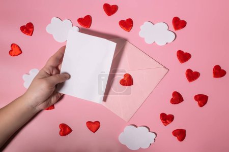 Hand hält ein leeres Buchstabenformular für Valentinstag oder Muttertag über einen Umschlag mit Herzen. Attrappe