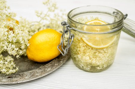 Foto de A jar of lemon and fresh elderberry inflorescences in syrup. Vintage metal plate on a white table. - Imagen libre de derechos