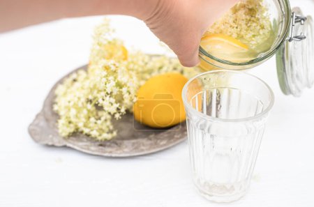 Foto de Hand pouring a refreshing summer drink of elderberry and lemon into a faceted glass on a white table. - Imagen libre de derechos