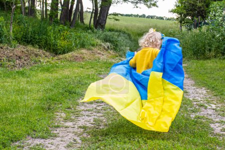 Foto de Niña pequeña con la bandera de Ucrania amarillo-azul en sus manos. niño corre en un camino de tierra en el campo contra el fondo de los árboles. bebé ríe felizmente y se divierte a pie. - Imagen libre de derechos