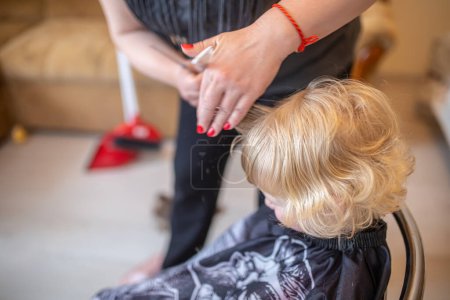 Photo for The hairdresser pulls out the hair on the bangs before cutting. A little fair-haired girl in a home does a hairstyle. Consept Beauty Salon - Royalty Free Image