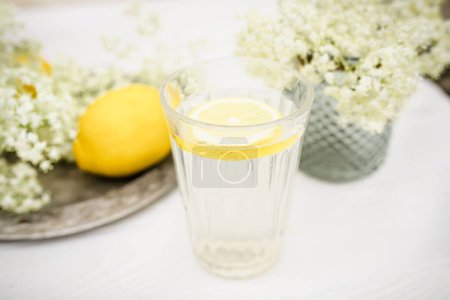 Foto de A faceted glass with a slice of lemon near a vase with fresh elderberry flowers on a white background. - Imagen libre de derechos
