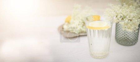 Foto de A faceted glass with a slice of lemon near a vase with fresh elderberry flowers on a white background. - Imagen libre de derechos
