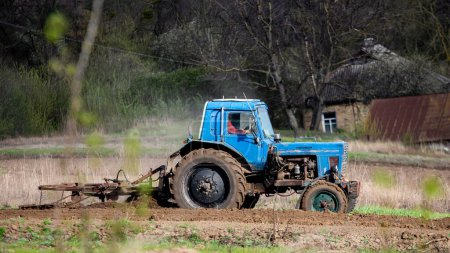 Téléchargez les photos : Vieux tracteur bleu avec charrue sur le champ et cultive la terre. Préparation du sol pour la plantation de légumes au printemps. Machines agricoles, travaux sur le terrain. - en image libre de droit