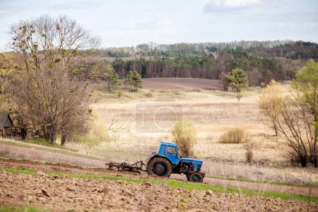 Photo for Old blue tractor with plow on field and cultivates soil on background of hilly landscape in Ukrainian village. Preparing the soil for planting vegetables in spring. Agricultural machinery, field work - Royalty Free Image