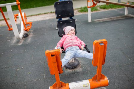Foto de Chica haciendo ejercicios en las máquinas de ejercicio al aire libre LEG PRESS, acostados en un sillón y descansando sus piernas. Equipo de fitness cardiovascular fácil de usar en el campo de deportes en el parque de la ciudad. - Imagen libre de derechos