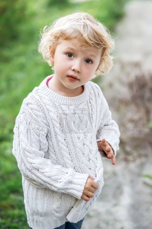 Téléchargez les photos : Portrait d'une petite fille blonde en plein air. Enfant en pull tricoté blanc sur fond d'herbe - en image libre de droit