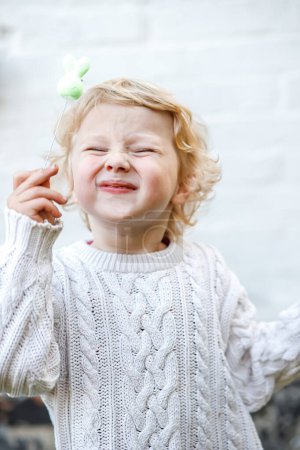 Photo for Portrait of a little girl squinting her eyes and holding a toy on a stick. Child in warm knitted sweater on white wall background outdoors - Royalty Free Image