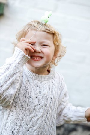 Foto de Retrato de una niña sonriendo y cubriéndose la cara con un juguete. Chica rubia en un suéter de punto blanco. - Imagen libre de derechos