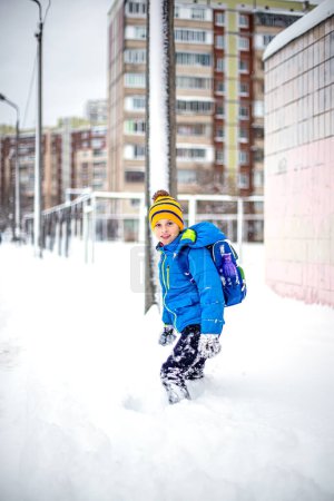 Téléchargez les photos : Adorable petit garçon qui s'amuse à l'extérieur par une belle journée d'hiver. Joyeux enfant jouant à l'extérieur. Un jeune étudiant rentre de l'école avec un sac à dos après une chute de neige en hiver. - en image libre de droit