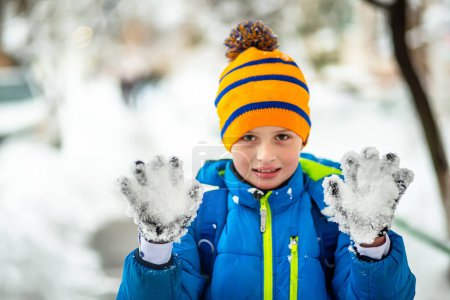 Foto de Niño estudiante de chaqueta azul mostrando palmas con guantes cubiertos de nieve durante las nevadas en la ciudad. Un niño divertido con ropa azul de invierno camina durante una nevada. Actividades al aire libre de invierno para niños. - Imagen libre de derechos