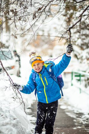 Foto de Niño estudiante con mochila en los hombros que se divierten en el camino nevado arrojando nieve de ramas de árboles. Un niño divertido con ropa azul de invierno camina durante una nevada. Actividades al aire libre de invierno para niños. - Imagen libre de derechos