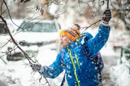 Foto de Chico divertido vertiendo nieve sobre sí mismo de las ramas de los árboles durante las nevadas en invierno mientras camina al aire libre. Un niño divertido con ropa azul de invierno camina durante una nevada. Actividades al aire libre de invierno para niños. - Imagen libre de derechos