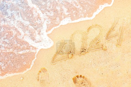 Téléchargez les photos : L'année à venir est 2024. Bonne année en bord de mer. La vague océanique emporte le sable près de l'inscription sur le sable sous forme de nombres 2024. - en image libre de droit